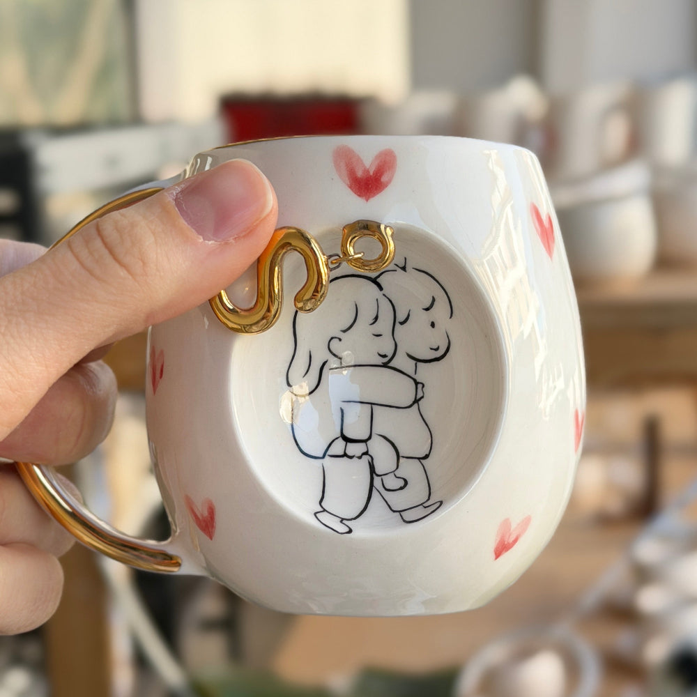 El yapımı harfli porselen kupa, beyaz zemin üzerinde el boyaması kalp desenleri ve 24K altın kaplama harf
